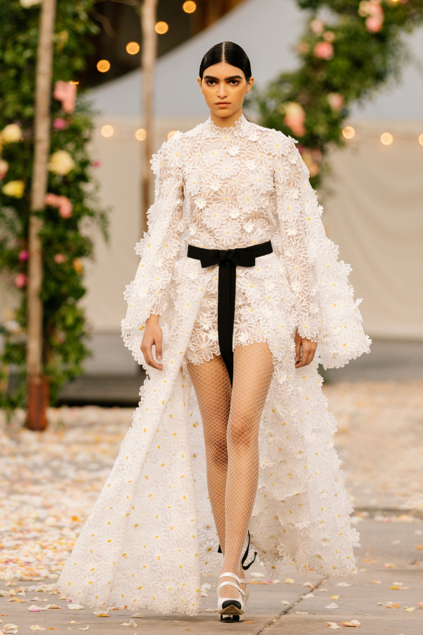 A long removable skirt in lace made of white daisies, that liberates the legs, is worn open over a dress in the same lace with an integrated cape.