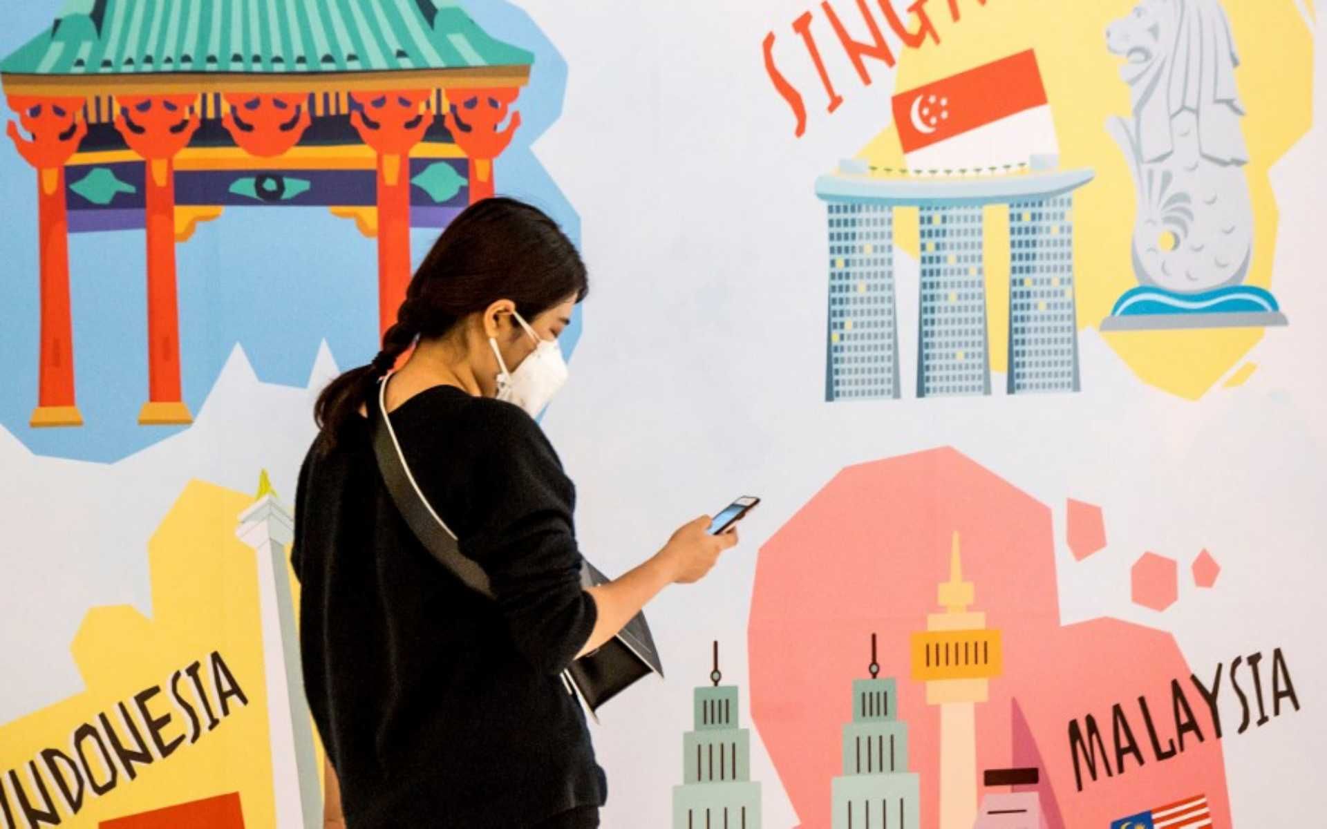 A woman, wearing a protective facemask amid fears over the spread of the COVID-19 coronavirus, walks in front of an advertisement board in Bangkok on February 17, 2020, featuring attractions from Japan, Singapore, Indonesia and Malaysia. (Photo by Mladen ANTONOV / AFP)