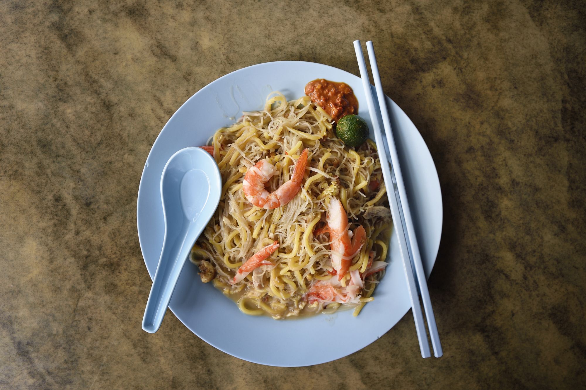 Singaporean style fried hokkien mee noodles with prawn, lime and sambal on a plastic plate with chopsticks and spoon