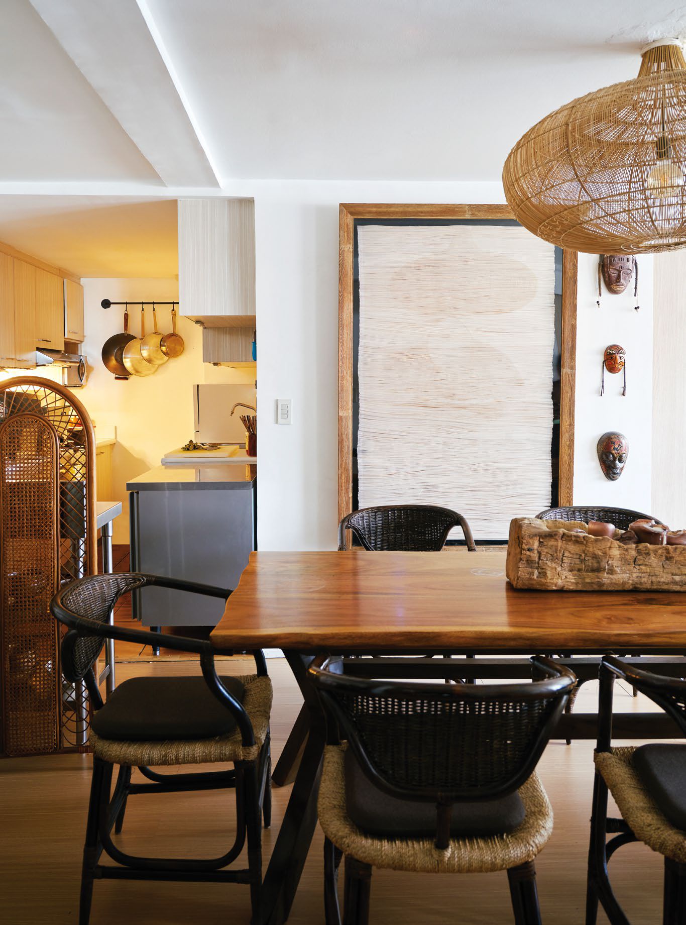 A wood slab dining table and rattan chairs with abaca seats are backdropped by a textile wall art by Olivia d’Aboville in the dining area
