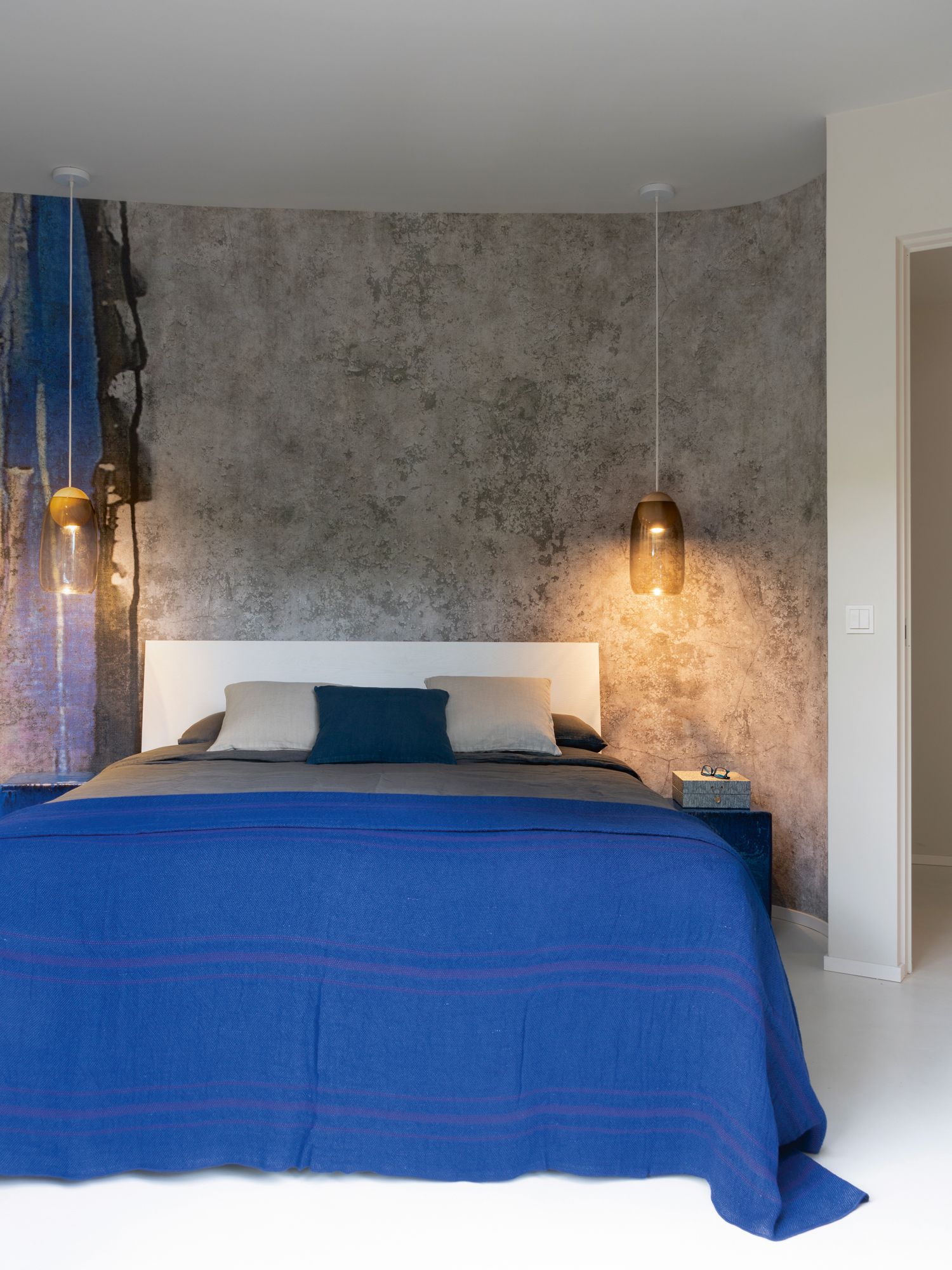 Vivid blue bedlinen and the concrete-like wallpaper from Glamora create a lively mix of colour and texture in this bedroom