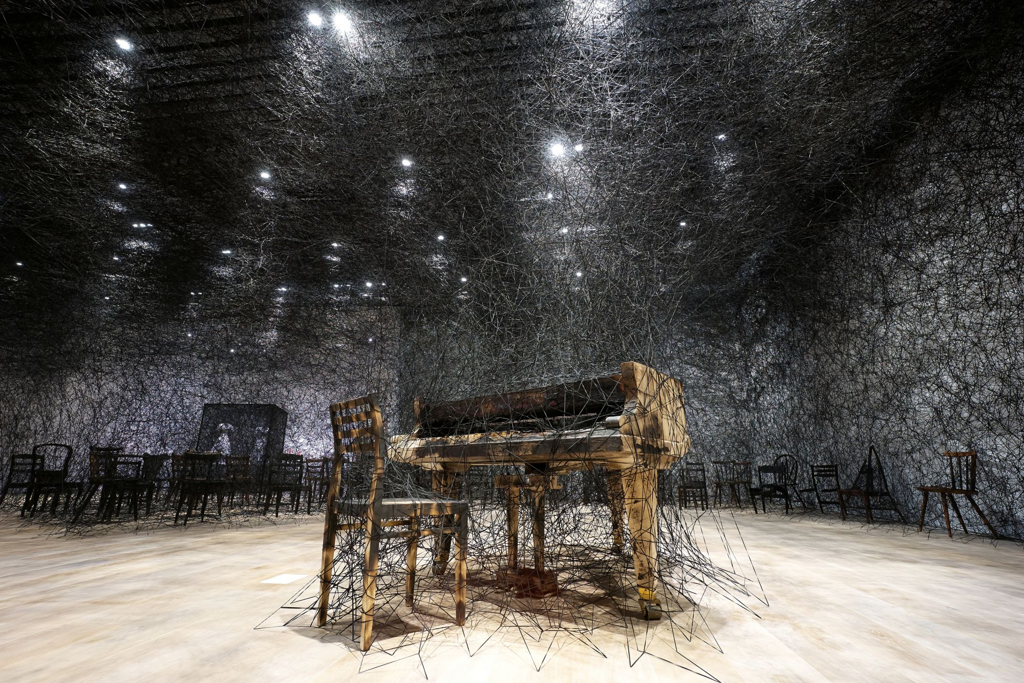 塩田千春,《寂靜中》(暫譯),2002/2019,燒焦鋼琴、燒焦椅、Alcantara黑線,尺寸視空間而定。 製作贊助:Alcantara S.p.A 由Kenji Taki 畫廊,名古屋/東京提供 展出現場:「塩田千春展:魂がふるえる」,東京森美術館,2019 攝影:Sunhi Mang 照片由森美術館提供