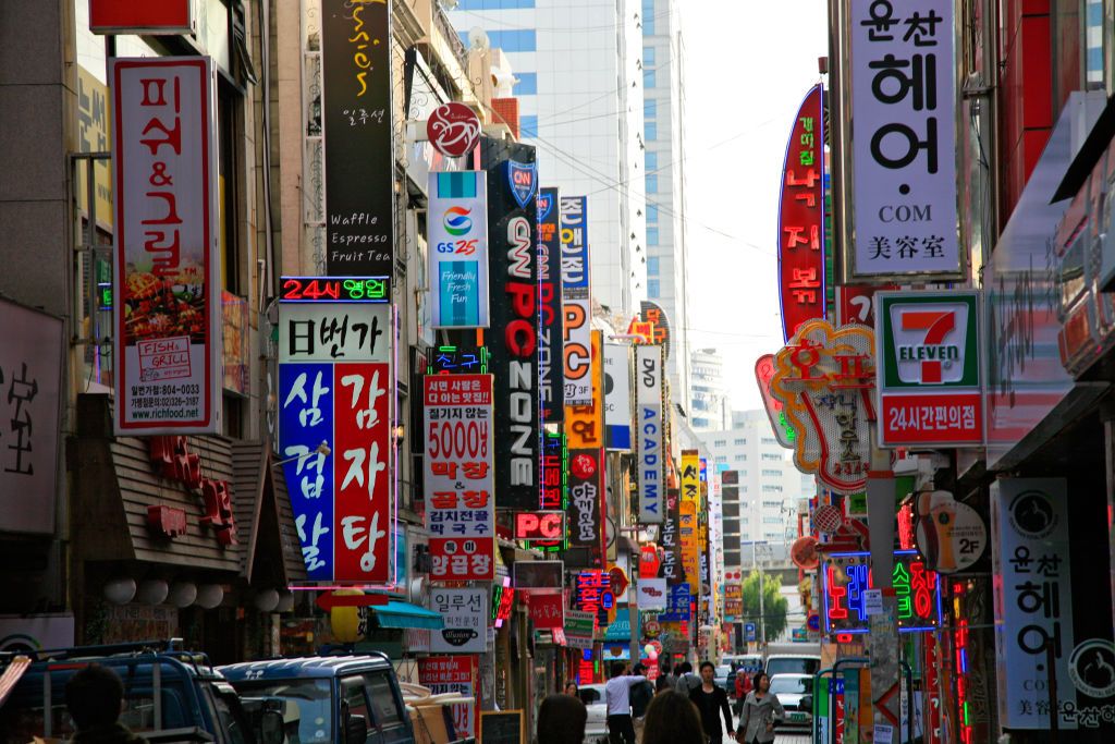 [UNVERIFIED CONTENT] Business advertisement street signs in Busan (Pusan), South Korea. Typical street with multitude of advertisements of various businesses including PC, computers, DVD, food, shops. Neon signs. Modern high-rise buildings on the background.
