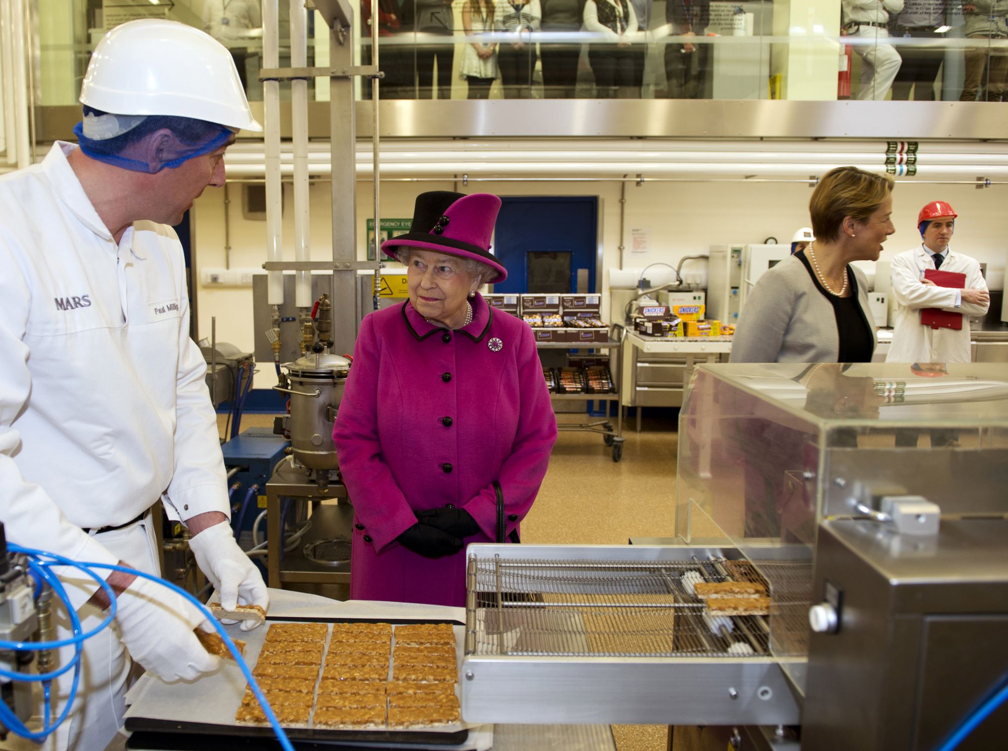 SLOUGH, UNITED KINGDOM - APRIL 05: Queen Elizabeth II looks on as Paul Milligan (L) puts Mars bars into a machine to cover them with chocolate in the pilot plant, part of the research and development, during a visit to Mars Chocolate UK in Slough on April 5, 2013. (Photo by Adrian Dennis - WPA Pool/Getty Images)