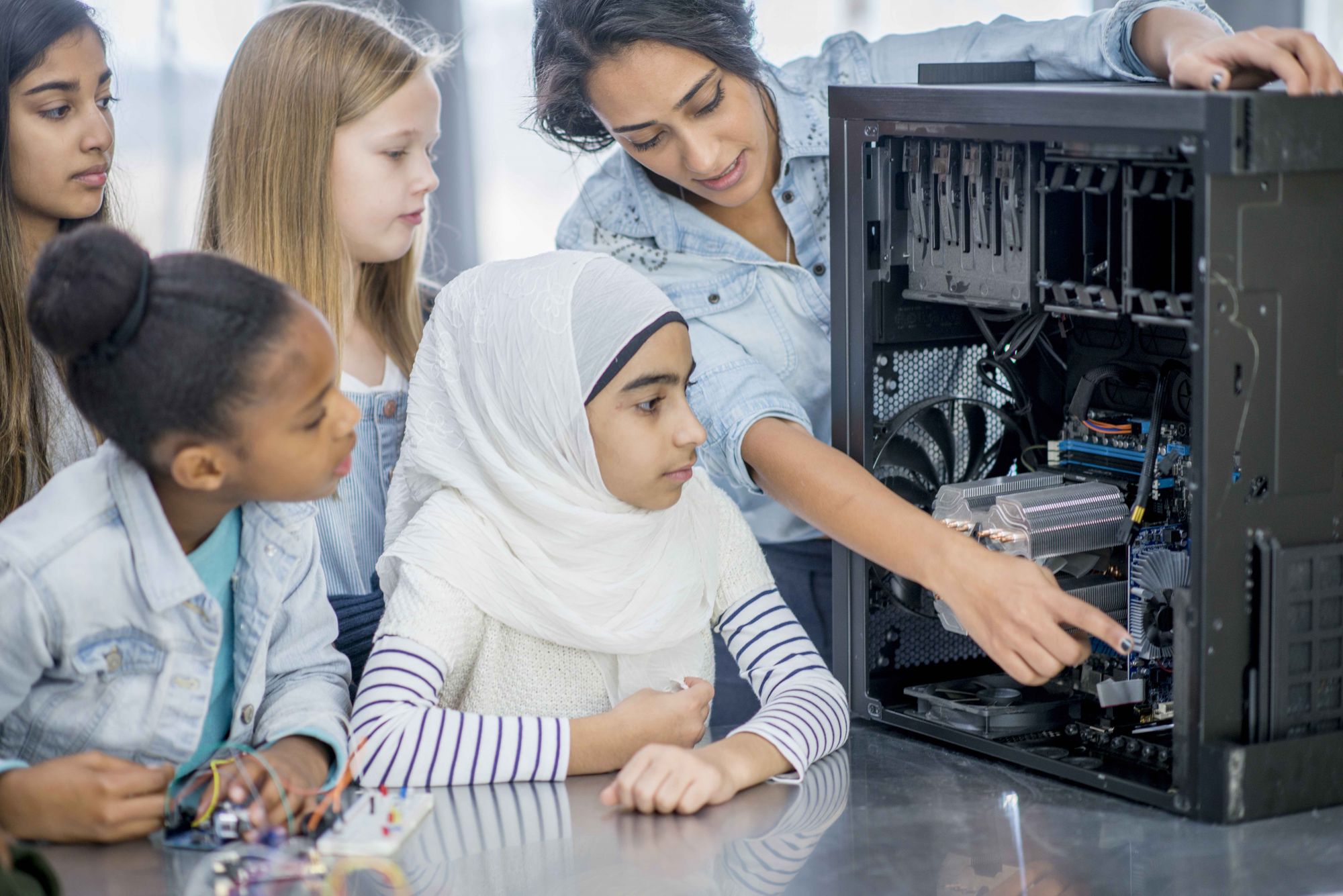 A group of elementary school children and their teacher and indoors in their classroom. The teacher has opened a desktop computer to view the inside components. She is pointing to certain parts and explaining what they do.