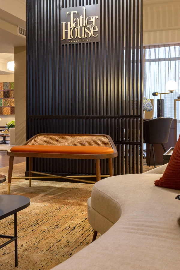 An elegant bench with a vibrant orange upholstered seat and woven back greets guests at Tatler House