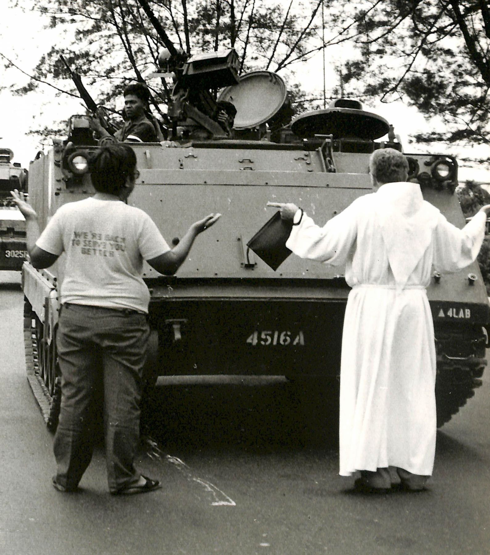 Two men hold their hands up in supplication as Marcos loyalists avoid looking at them. The priest had been facing them alone, until another man joined them ┃Photo: Presidential Museum And Library on Flickr
