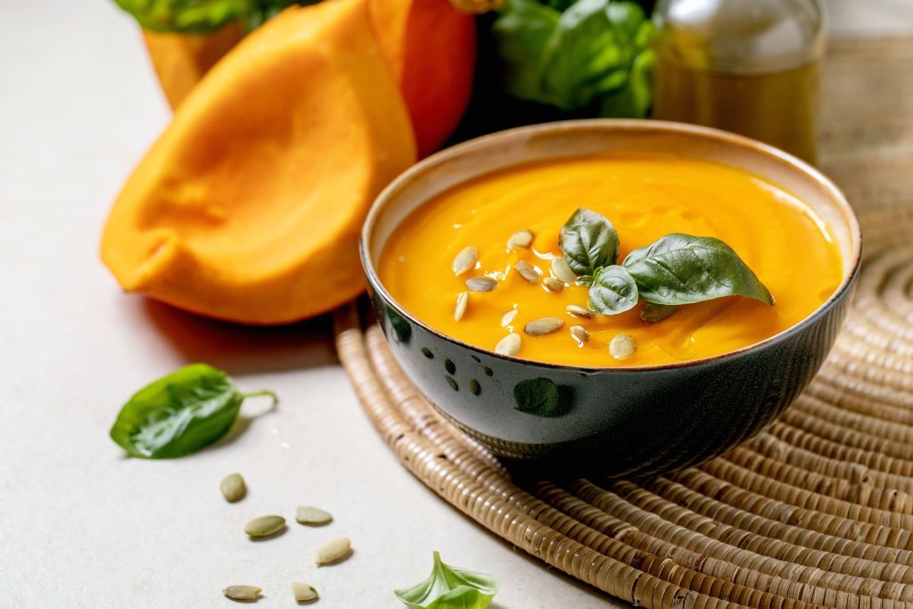 Bowl of pumpkin or carrot vegetarian cream soup decorated by fresh basil, olive oil and pumpkin seeds on white texture table with ingredients above. (Photo by: Natasha Breen/REDA&CO/Universal Images Group via Getty Images)