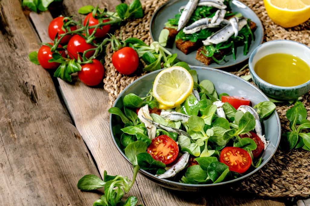 Green field salad with pickled anchovies or sardines fillet, and cherry tomatoes, served in blue bowl with lemon and olive oil on straw napkin over old wooden table.. (Photo by: Natasha Breen/REDA&CO/Universal Images Group via Getty Images)