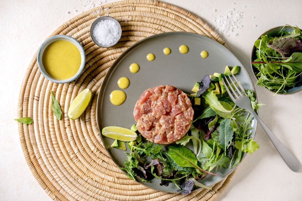 Tuna tartare with green salad. lime. avocado and mustard sauce serving on ceramic plate on straw napkin over white texture background. Flat lay. copy space. Fine dining. restaurant appetizer. (Photo by: Natasha Breen/REDA&CO/Universal Images Group via Getty Images)