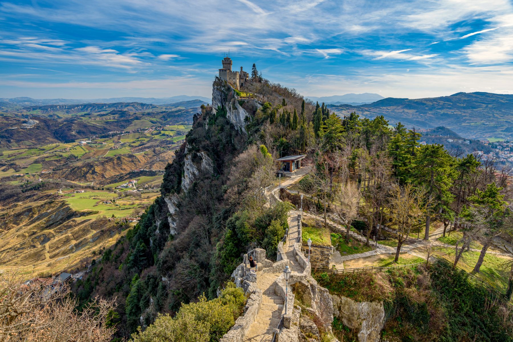 In San Marino there are 3 towers on the mountain ridge in a row. This is Falesia the second tower. The sun is shining and the sky is blue with white fluffy clouds.
