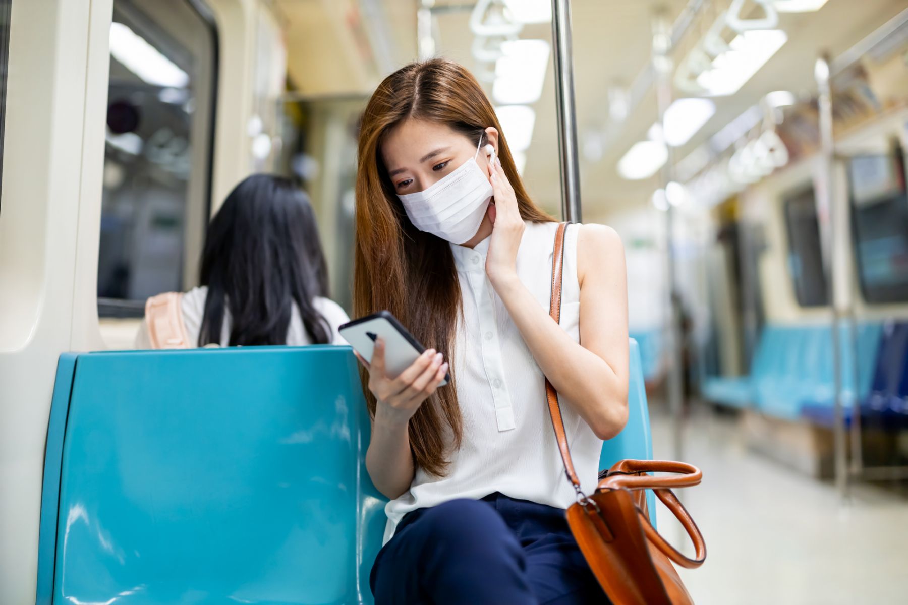 Asian woman use a smartphone with ear buds wearing surgical mask face protection and keep social distance while commuting in the metro or train