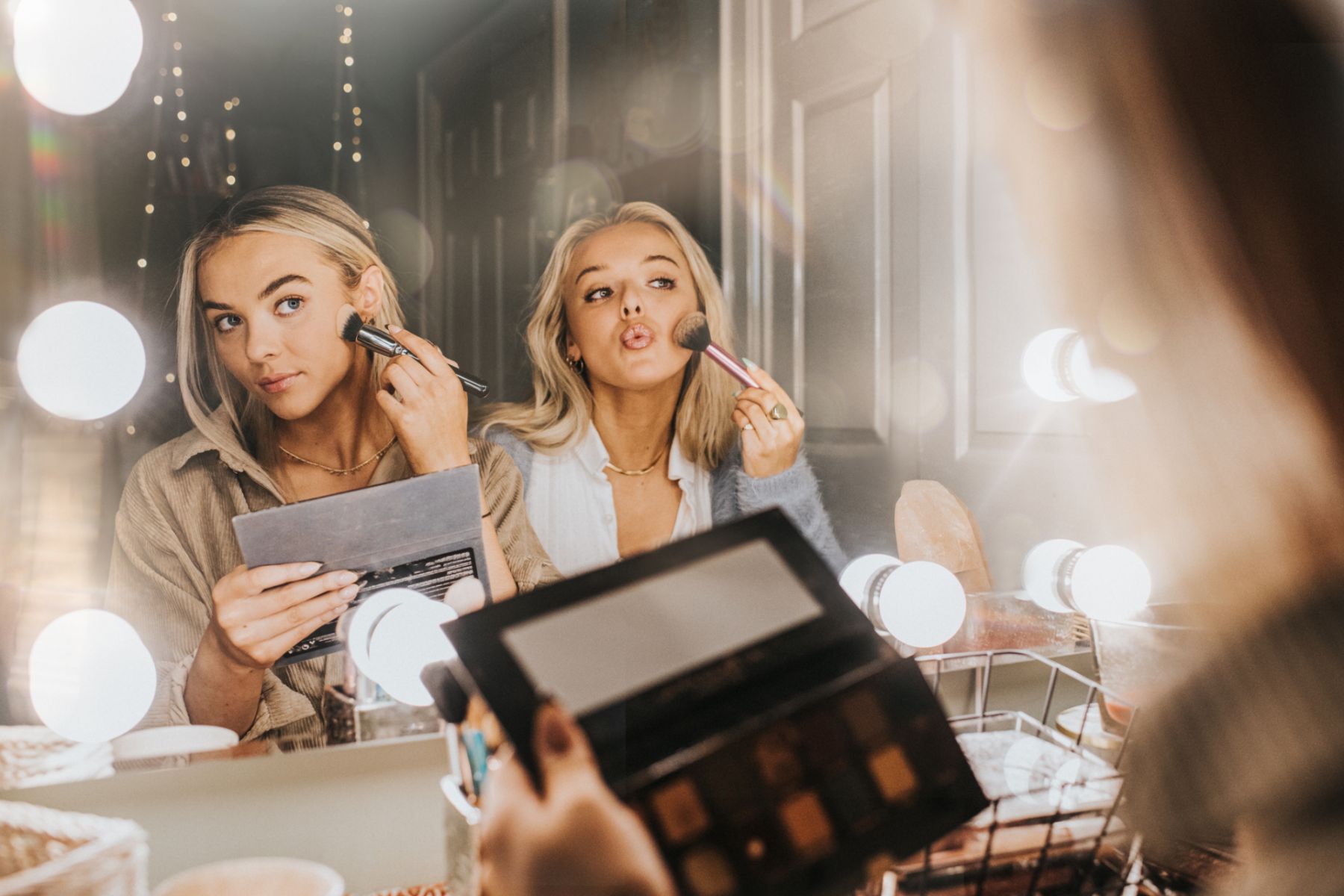 Two young woman in their twenties sit at a vanity table by a mirror surrounded by bulbs. They concentrate as they apply make-up to their faces with brushes. They are comfortable with each other. The scene is carefree and youthful as the lights create flare and reflections.