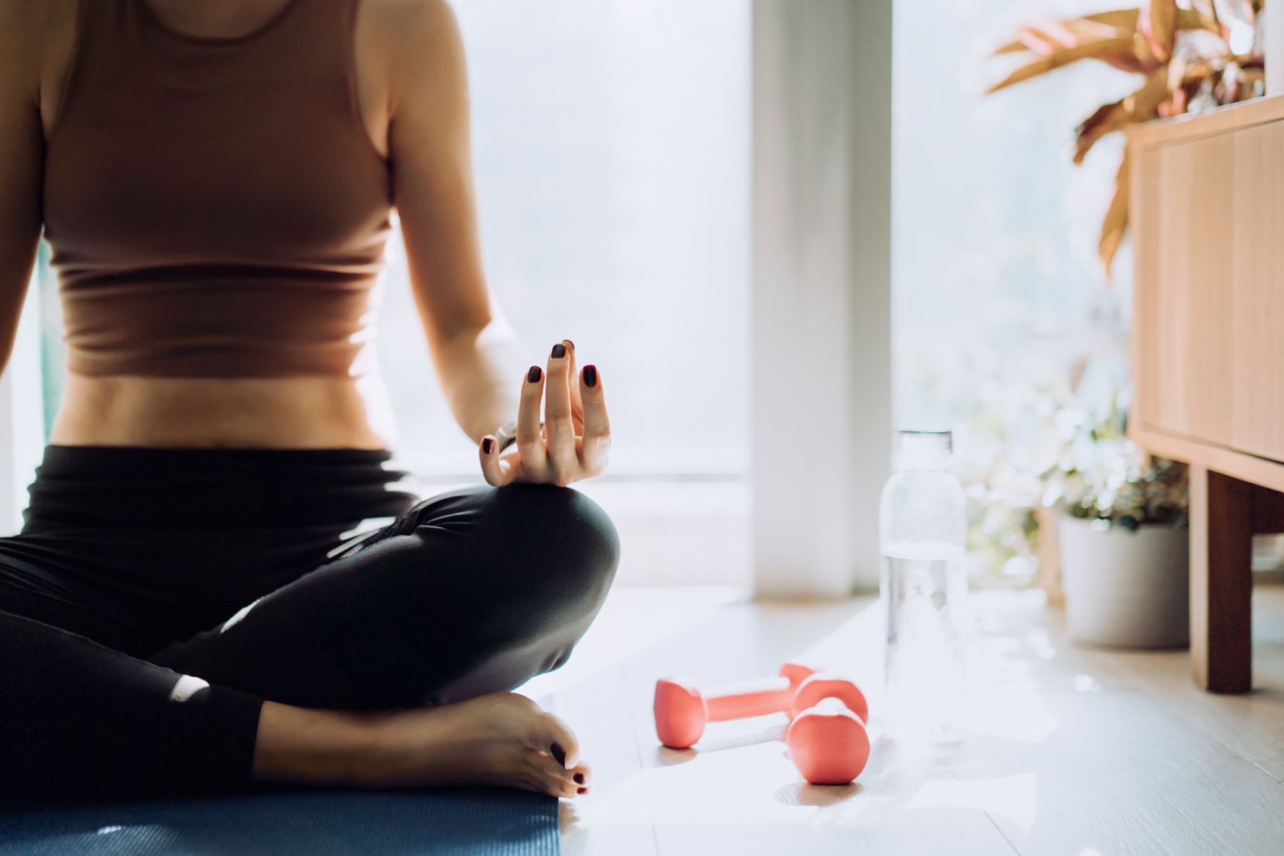 Cropped shot of young Asian woman sitting on the exercise mat, meditating in the lotus pose during a yoga session at home in the fresh bright morning. Fitness, wellness and home work out concept