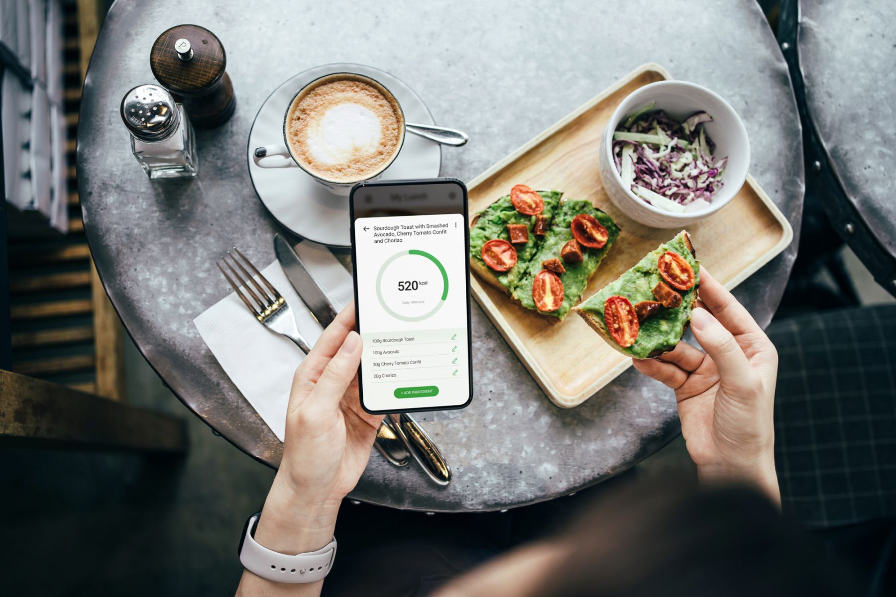 Overhead view of young Asian woman using fitness plan mobile app on smartphone to tailor make her daily diet meal plan, checking the nutrition facts and calories intake of her meal, sourdough toast with smashed avocado and cherry tomatoes in a restaurant