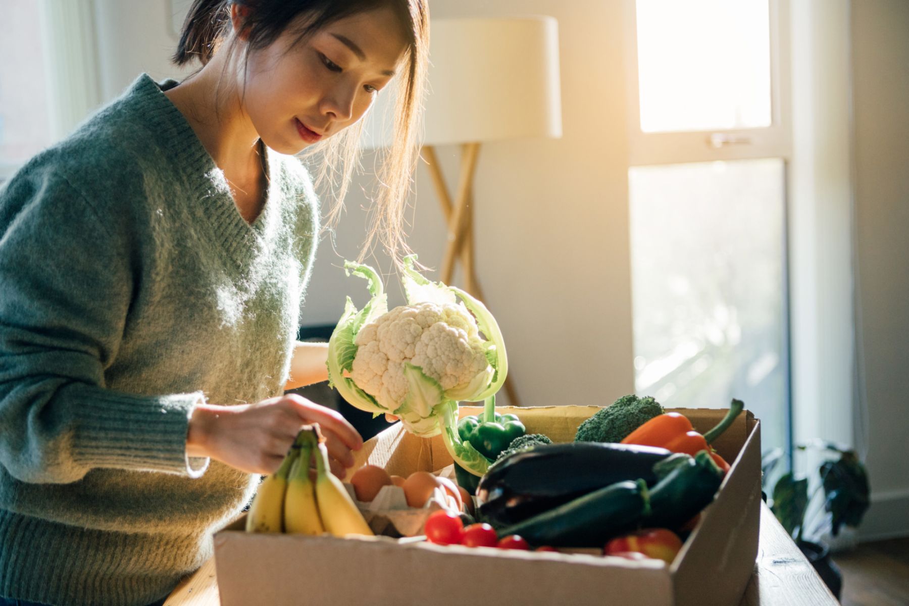 Beautiful smiling young woman picking up fresh organic vegetables from a delivery box to prepare cooking at home.