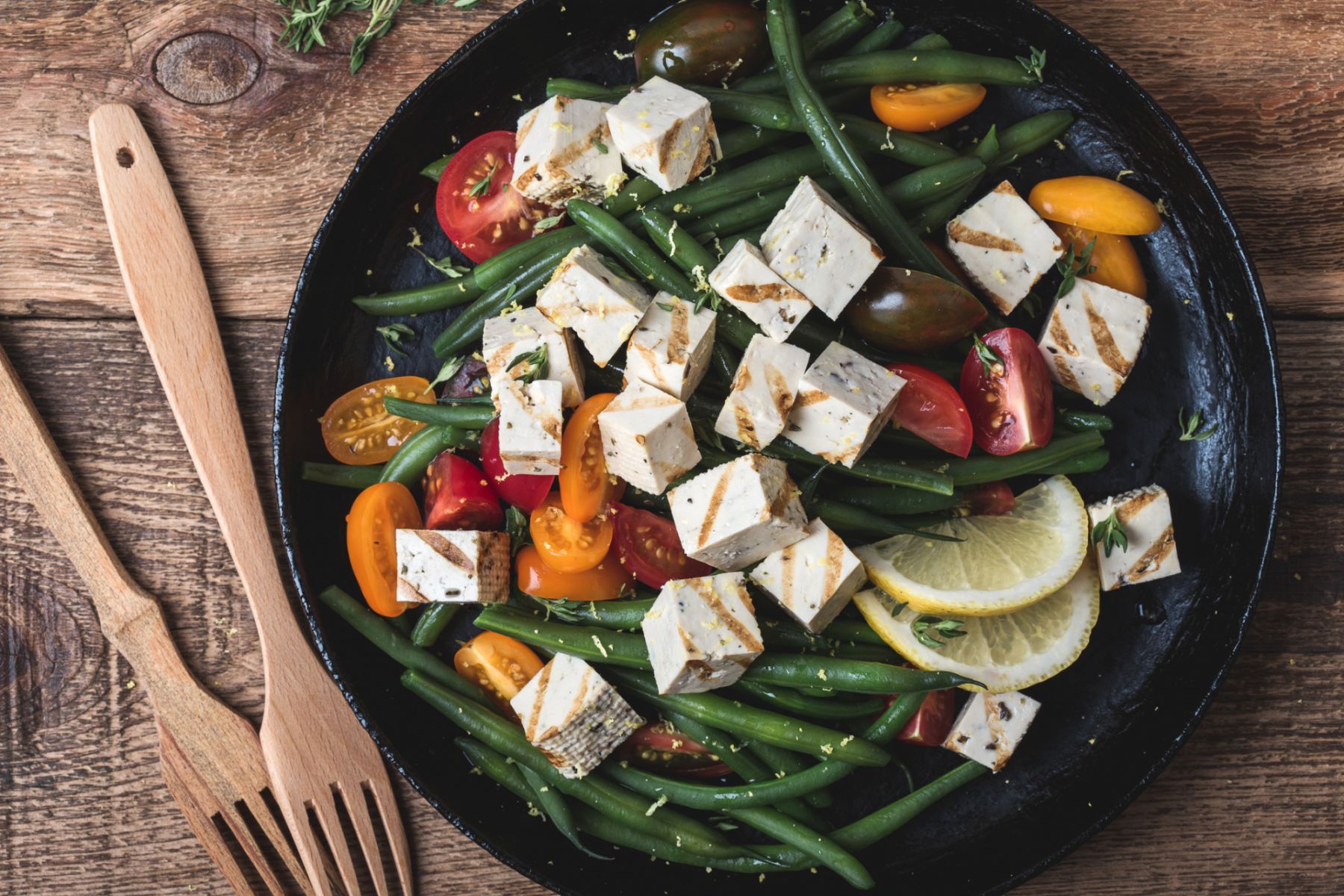Delicious summer vegan meal, healthy green beans salad with grilled tofu, fresh colorful mix cherry tomatoes, thyme herbs and lemon zest served in rural cast iron skillet, wooden forks, top view