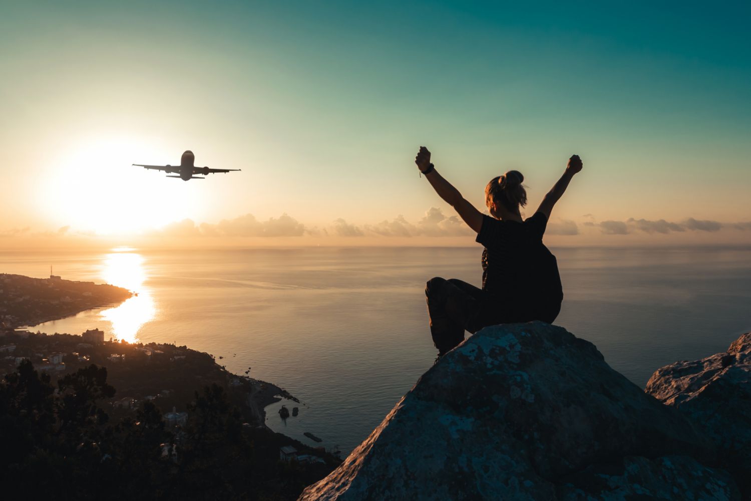 A teenage girl with a backpack sits on the edge of the peak of mountain with arms raised and watches an airplane flying over the sea coast at sunrise.