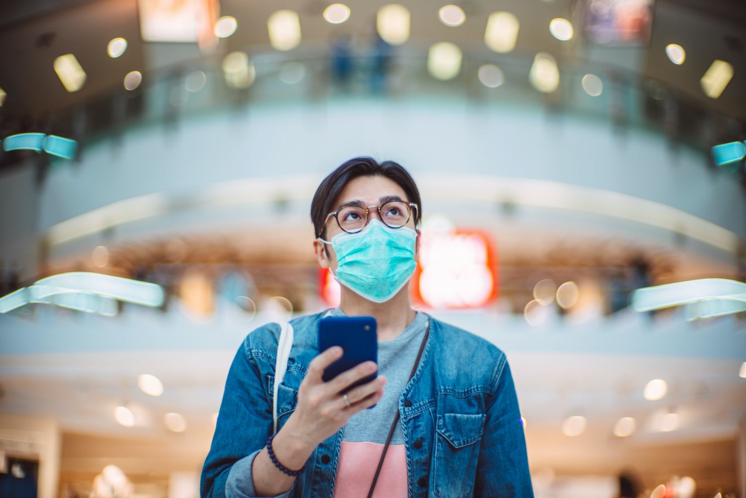 Young handsome Asian man in medical face mask using smartphone joyfully in shopping mall.