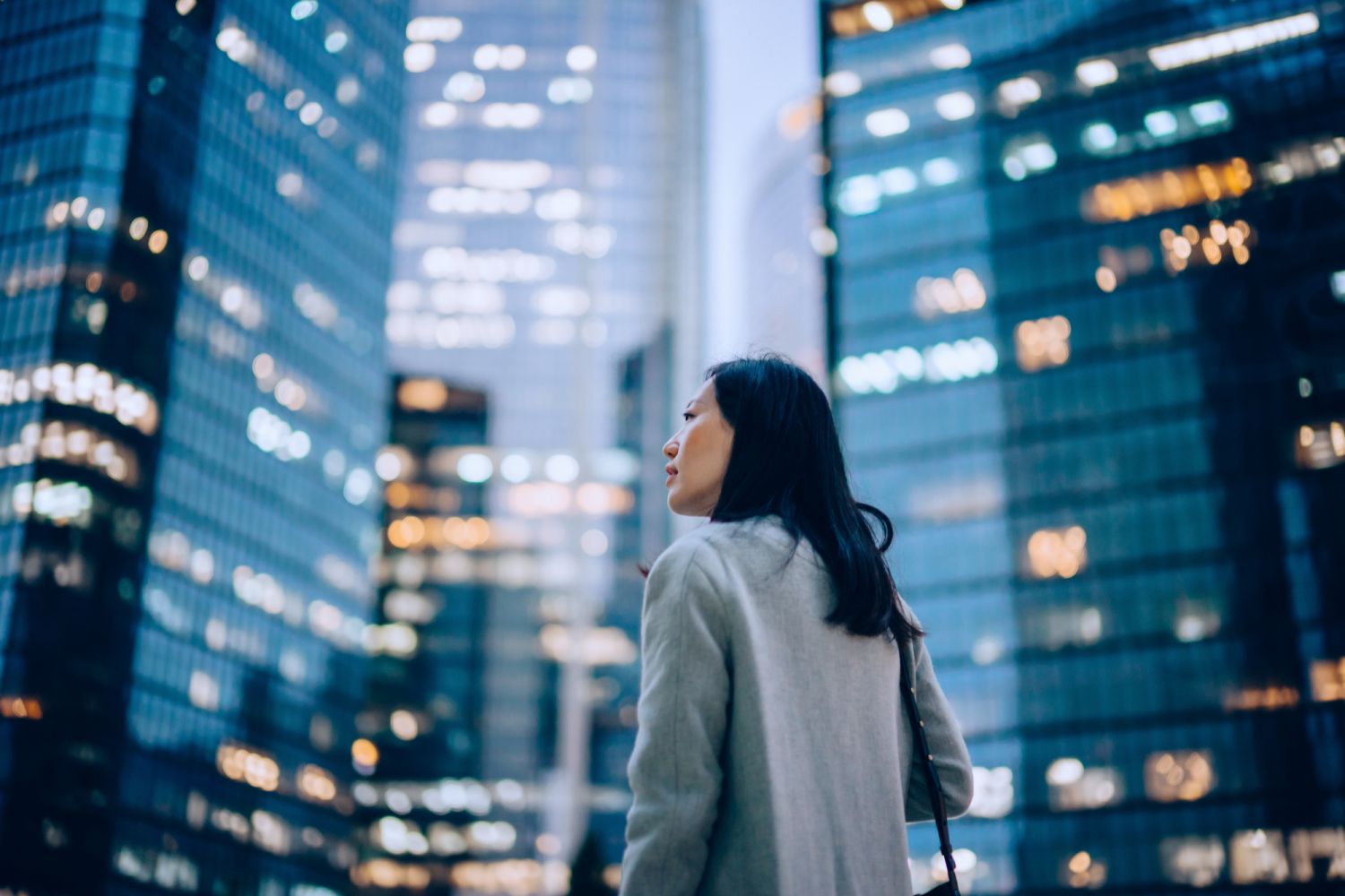 Low angle side profile of confident and professional young Asian businesswoman looking up while standing against contemporary corporate skyscrapers with illuminated facade in financial district in the evening. Female leadership and determined to success