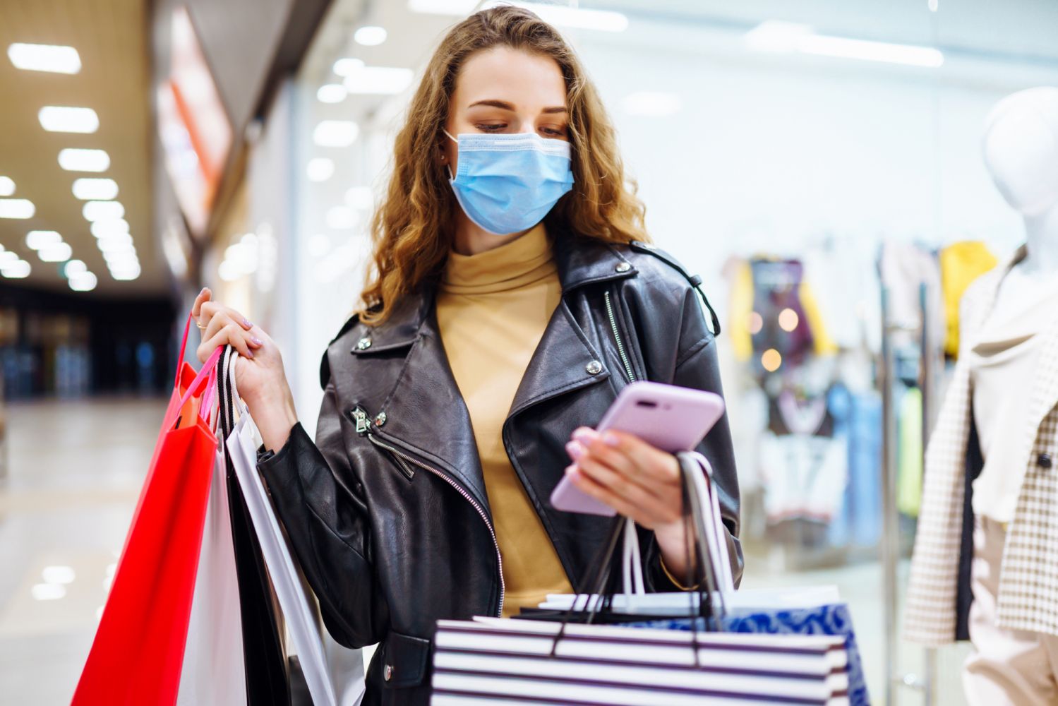 Fashionable  woman in protective medical mask with multi-colored  bags in modern shop center. Young woman using her phone. The joy of consumption, Gift shopping, holiday. Online shopping concept.
