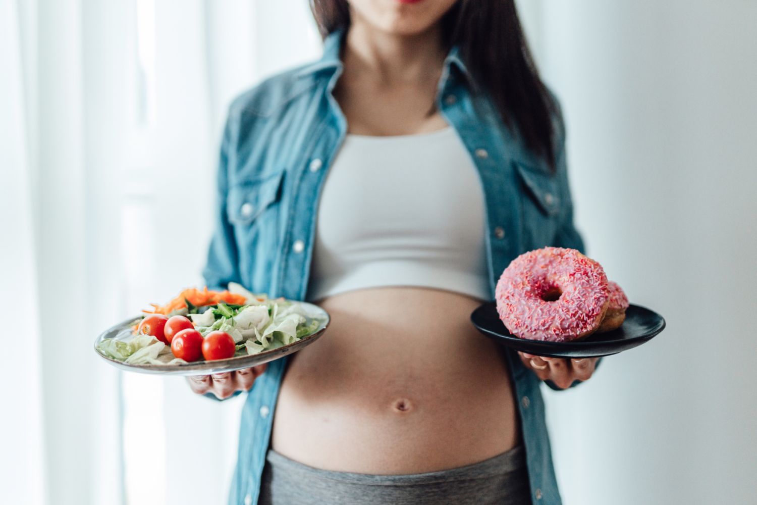 Midsection of pregnant woman choosing between vegetable saiad and doughnuts. Eating well in pregnancy. Food craving during pregnancy concept.