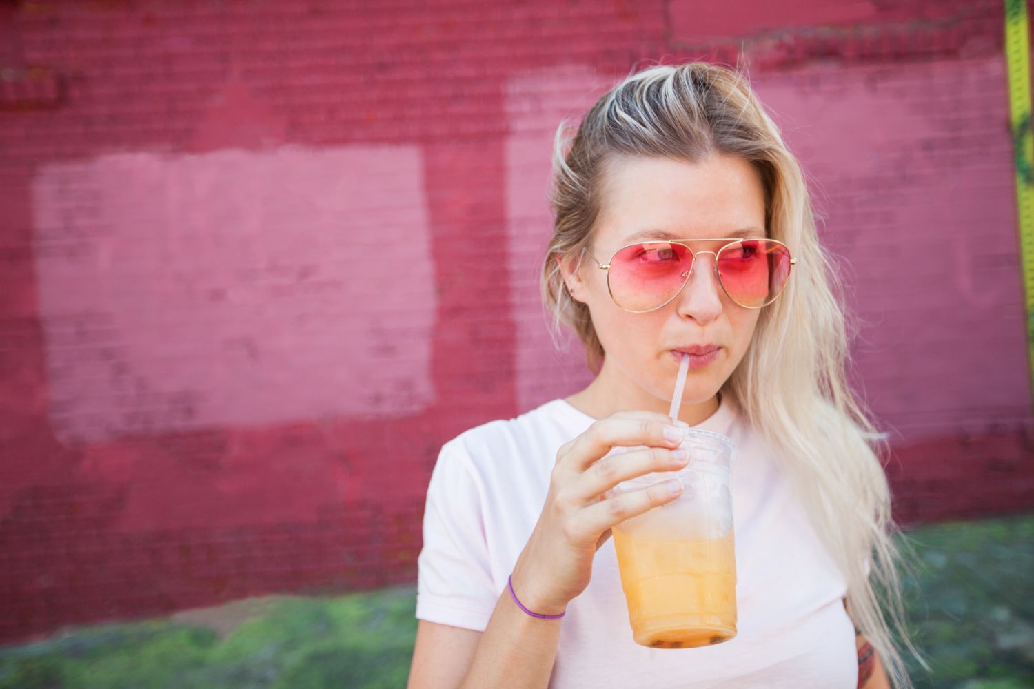 Young blonde woman wearing pink sunglasses drinking juice from a a plastic cup and straw in front of a pink wall