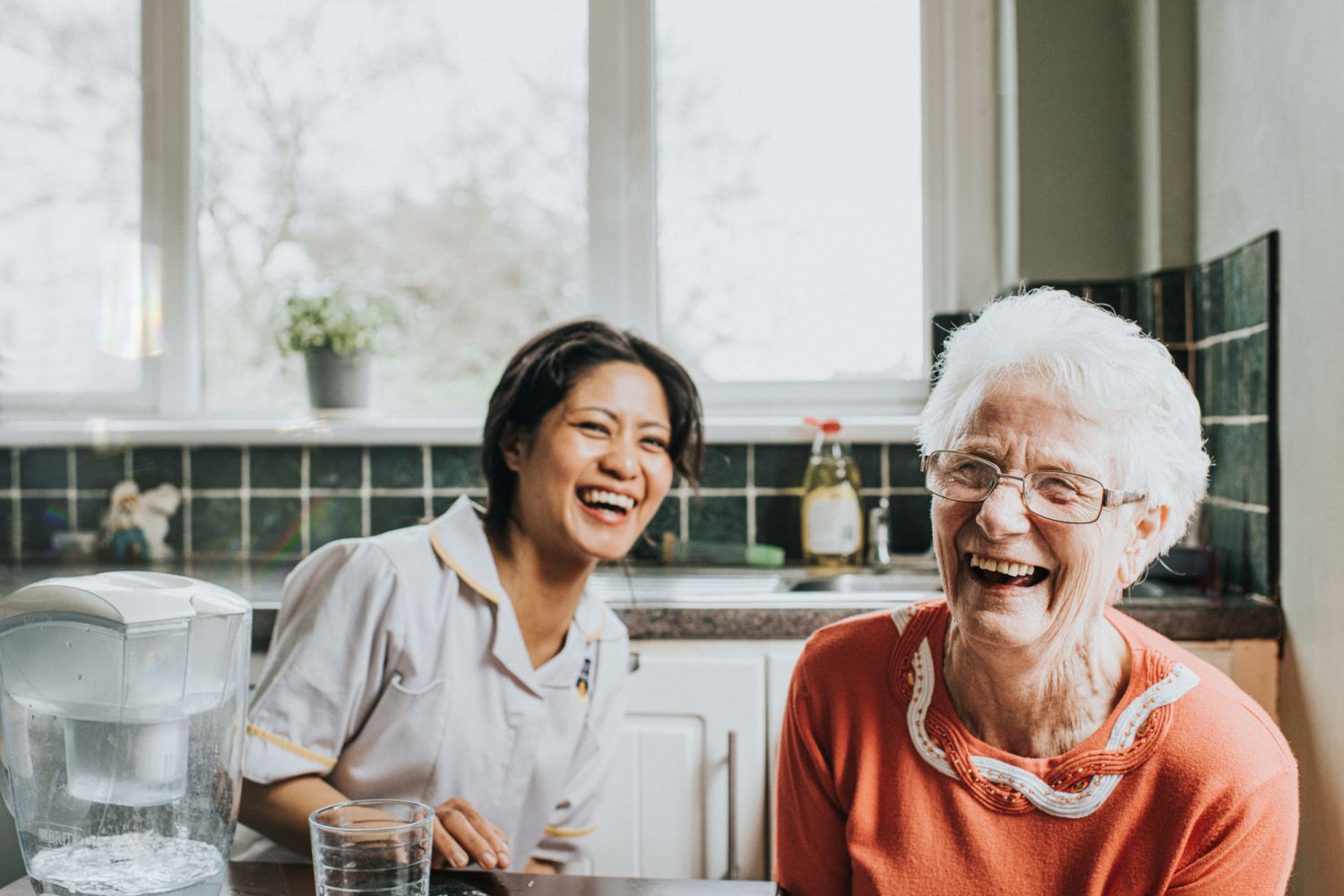 A young, friendly and bubbly carer sits beside an elderly woman at a kitchen table. The older woman faces the camera directly and laughs as the younger woman laughs behind her. The scene is lighthearted and fun, and the older woman quite clearly has a big personality.
