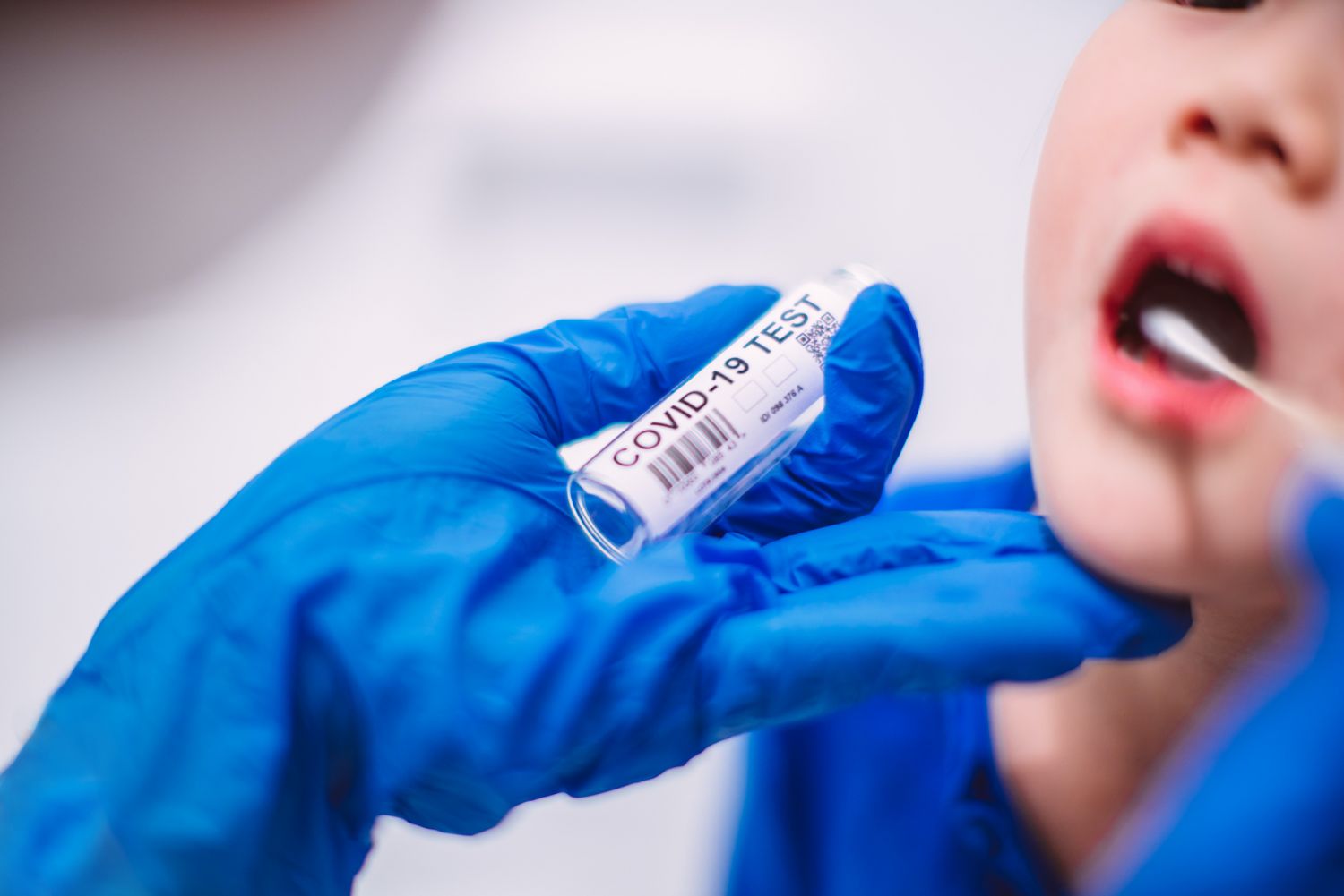 Doctorâs hands in protection gloves putting COVID-19 test swab into kidâs mouth in hospital.
