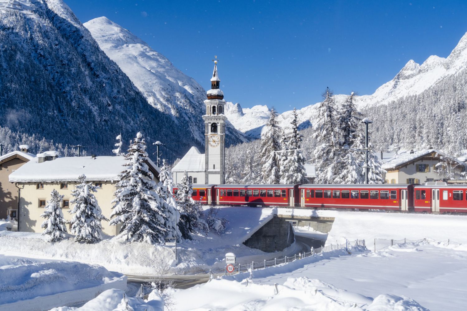 The famous red Bernina Express train passing by the bell tower of Bever alpine village covered with snow, Graubunden canton, Switzerland