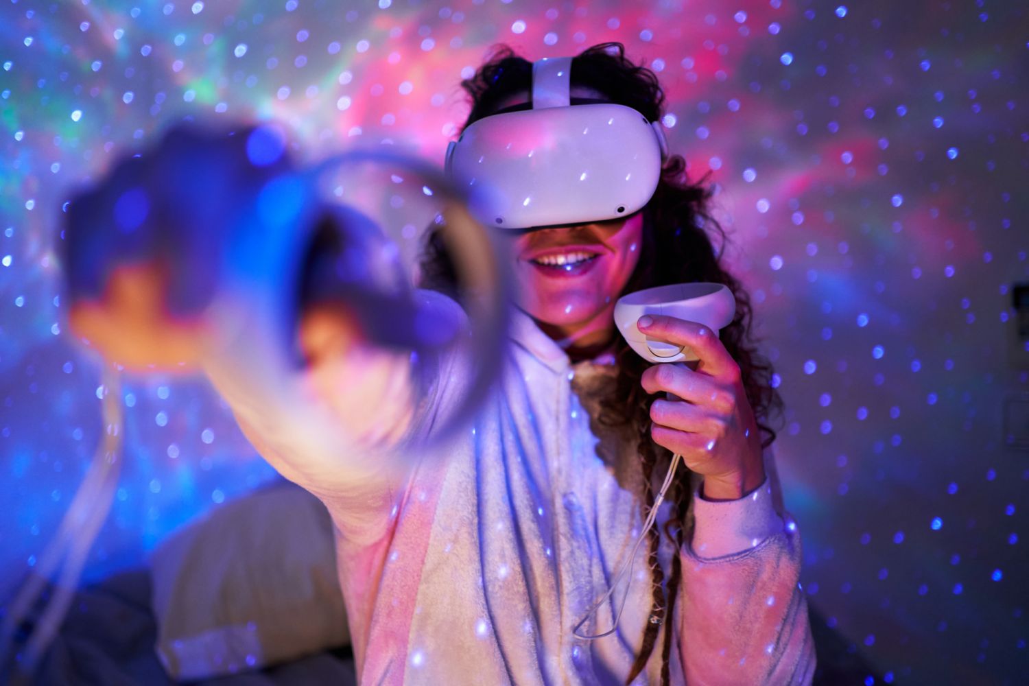 Cheerful young lady with long dark hair in warm pajama smiling and using modern controllers while exploring virtual reality in headset in illuminated dark room