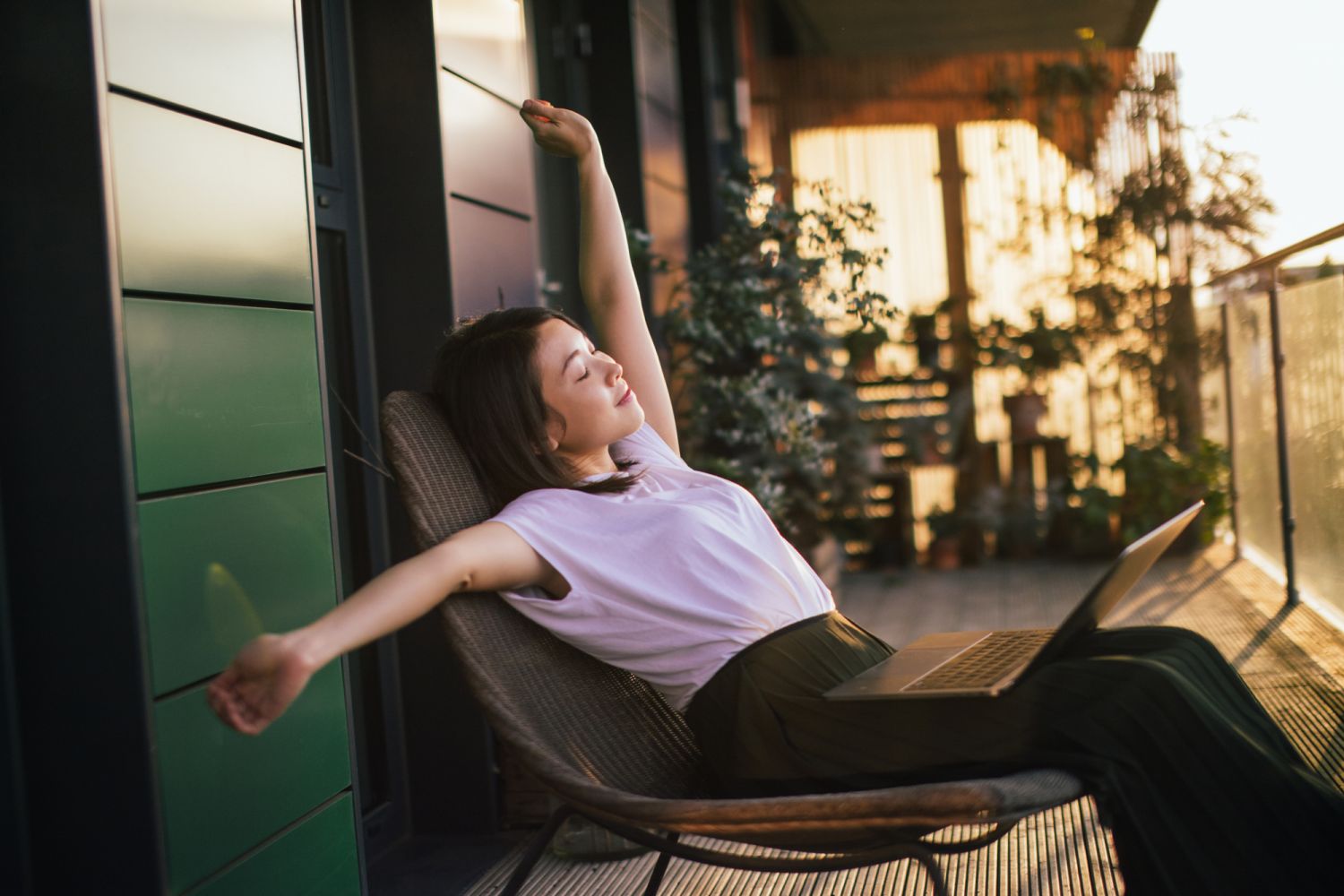 Young beautiful Asian woman stretching arms while working with laptop, sitting at the balcony. Remote working concept. Work-life balance concept.