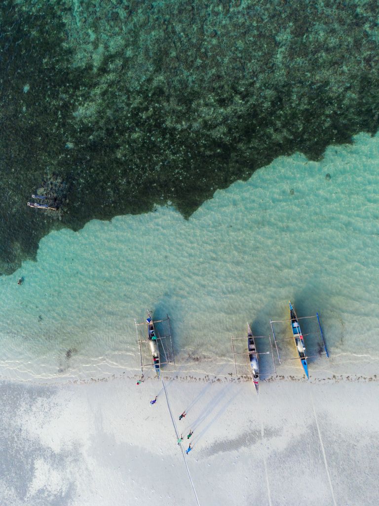 A drone photo of three outrigger canoes being brought out to sea at the shore of Bantayan Island of the coast of Cebu in the Philippines. The boats are lying at the shore of the crystal clear sea while a group of men is walking back on the beach to collect the next one. The sea floor can be seen in the crystal clear waters and the men cast long shadows on the beach