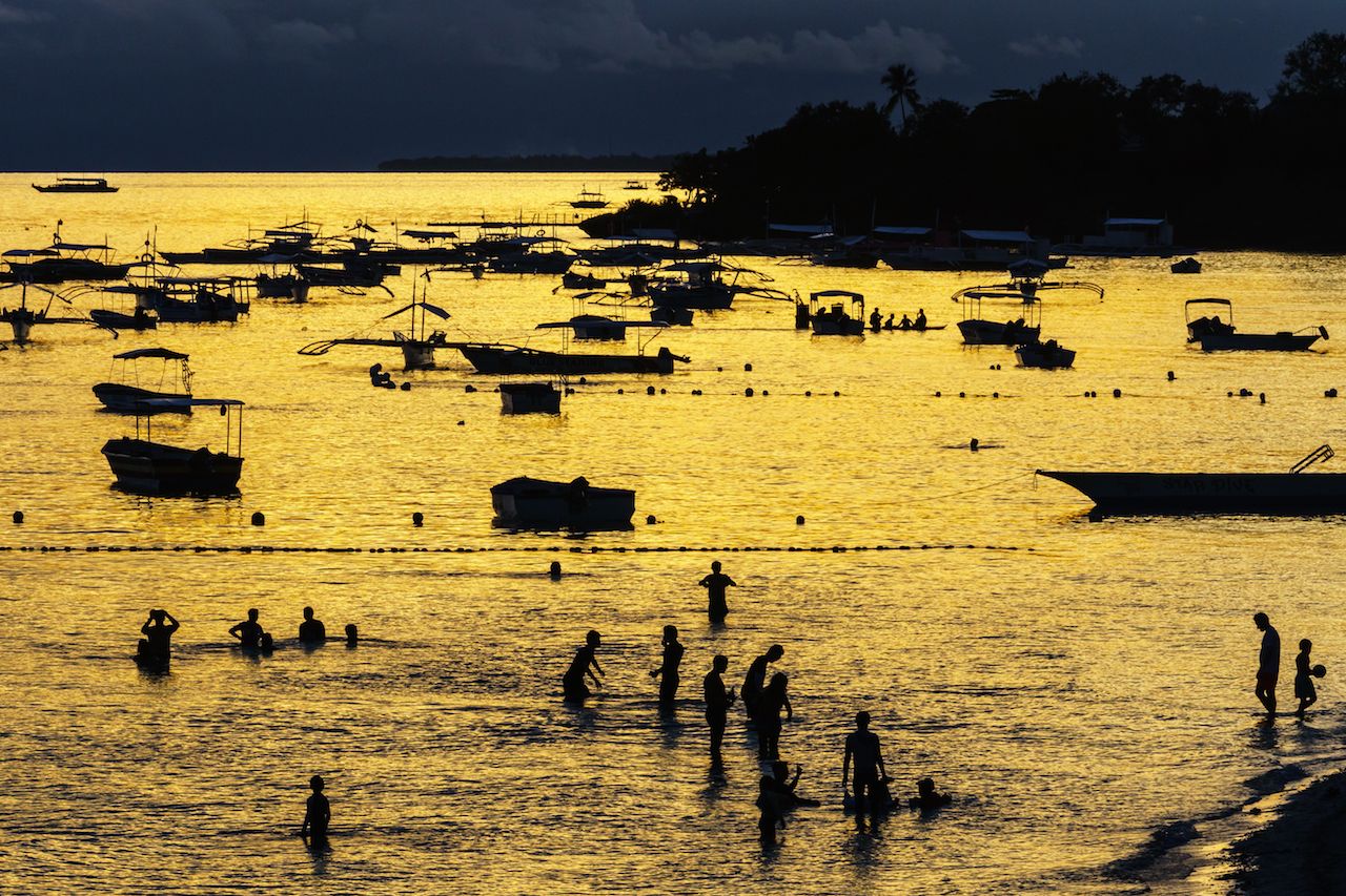 Golden light bathes the sea and highlights the silhouettes of boats and evening bathers, Panglao Beach, Bohol Island, Philippines