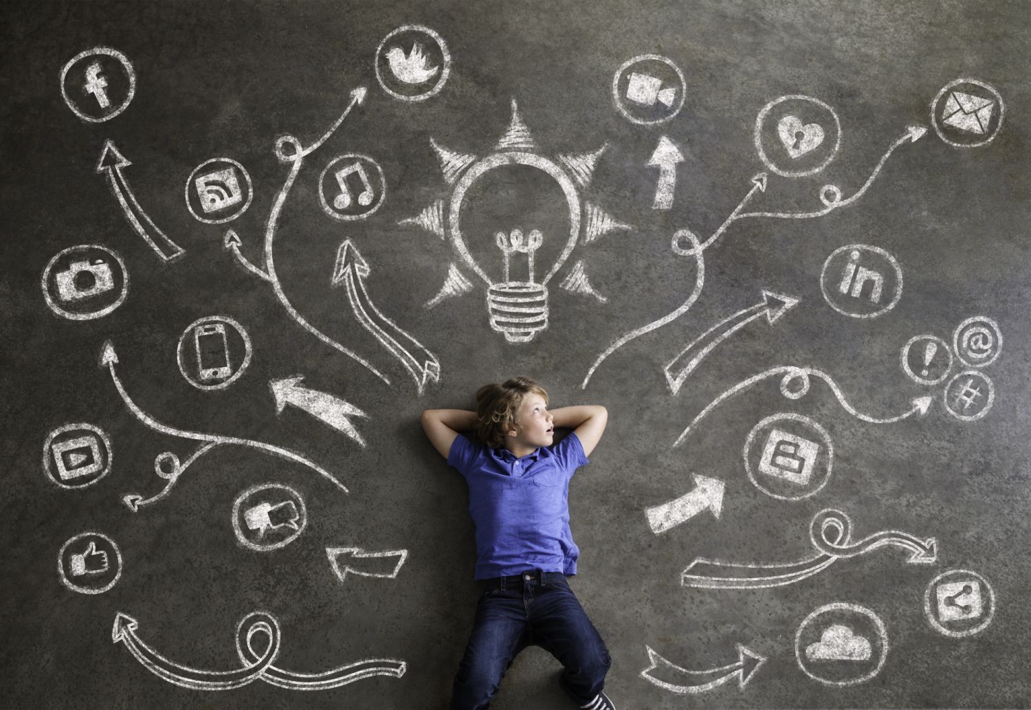 Elementary school boy lays inside a universe of chalkboard social media, music, communications, tech symbols