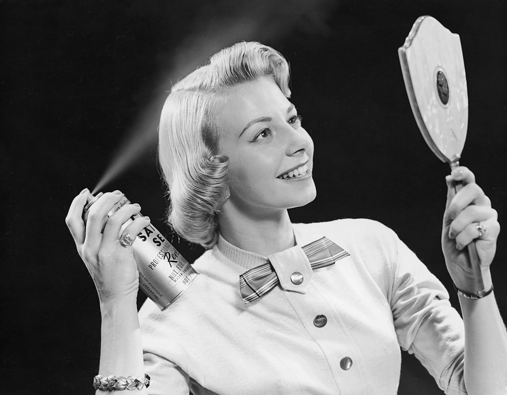 circa 1955:  A young woman uses a hand-held mirror to apply Revlon hair spray to her hair.  (Photo by Harold M. Lambert/Lambert/Getty Images)