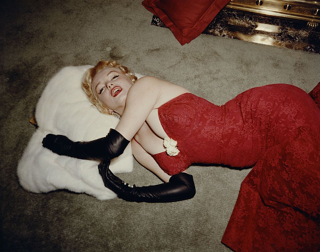 1955: EXCLUSIVE American actor Marilyn Monroe (1926 - 1962), wearing a red brocade evening gown and long black gloves, lying on a carpet and using a white fur stole as a pillow. (Photo by Gene Lester/Getty Images)
