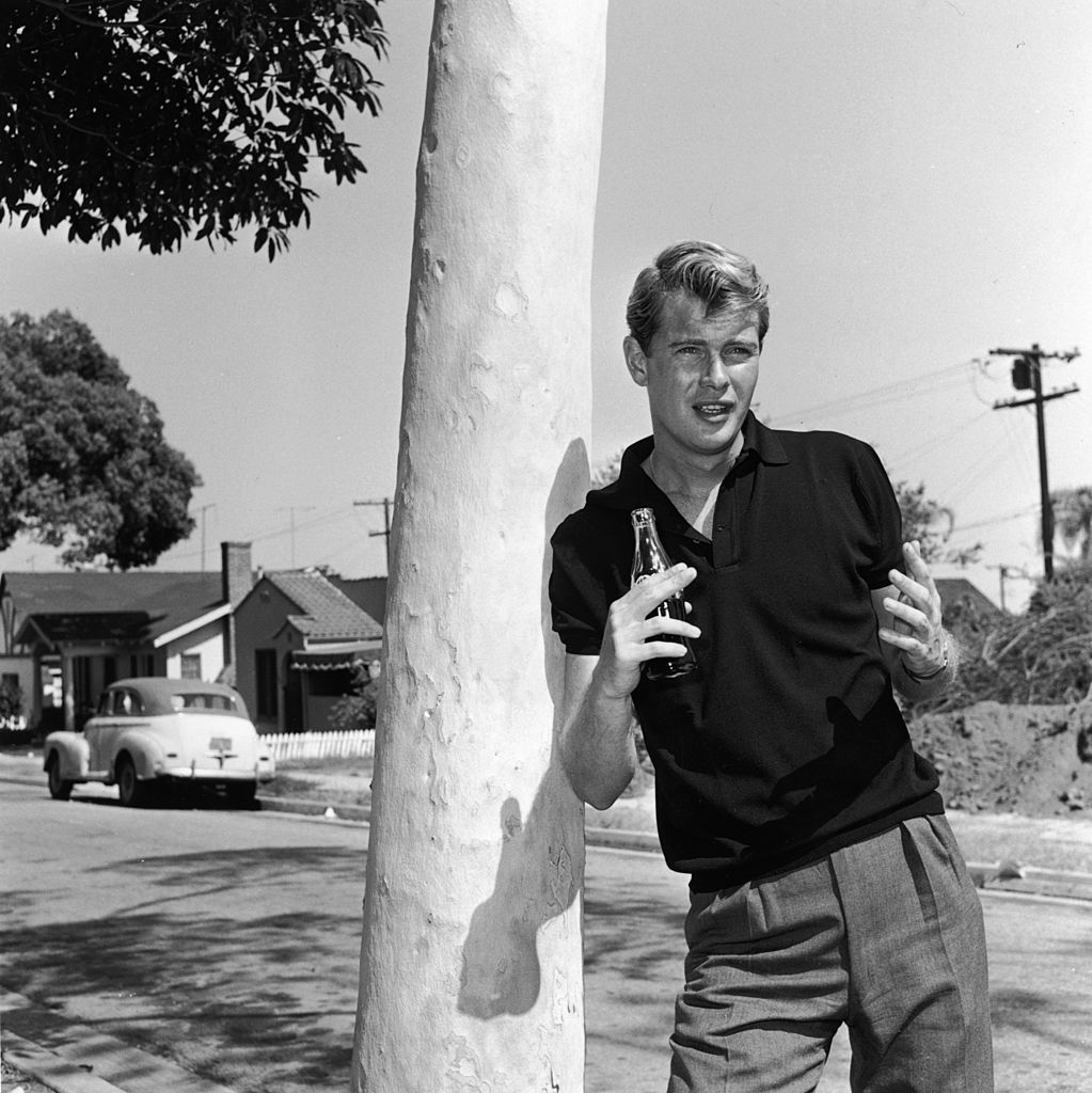 1956: Portrait of American actor Troy Donahue (1936 - 2001) leaning against a tree and gesturing with his hands while holding a Coca-Cola bottle on a residential street, California. Donahue wears a dark polo shirt and pleated pants. (Photo by Hulton Archive/Getty Images)