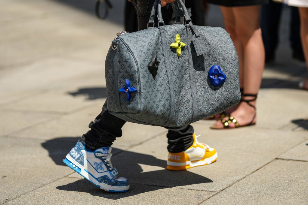 PARIS, FRANCE - JUNE 23: A guest wears a white t-shirt, a gray and blue denim LV monogram print pattern jacket from Louis Vuitton, black denim large pants, a gray leather embossed LV monogram pattern Keep All handbag from Louis Vuitton with embroidered large logo, a blue and white LV monogram print pattern sneakers from Louis Vuitton, a matching orange and white LV monogram print pattern sneakers from Louis Vuitton, outside the Louis Vuitton show, during Paris Fashion Week - Menswear Spring/Summer 2023, on