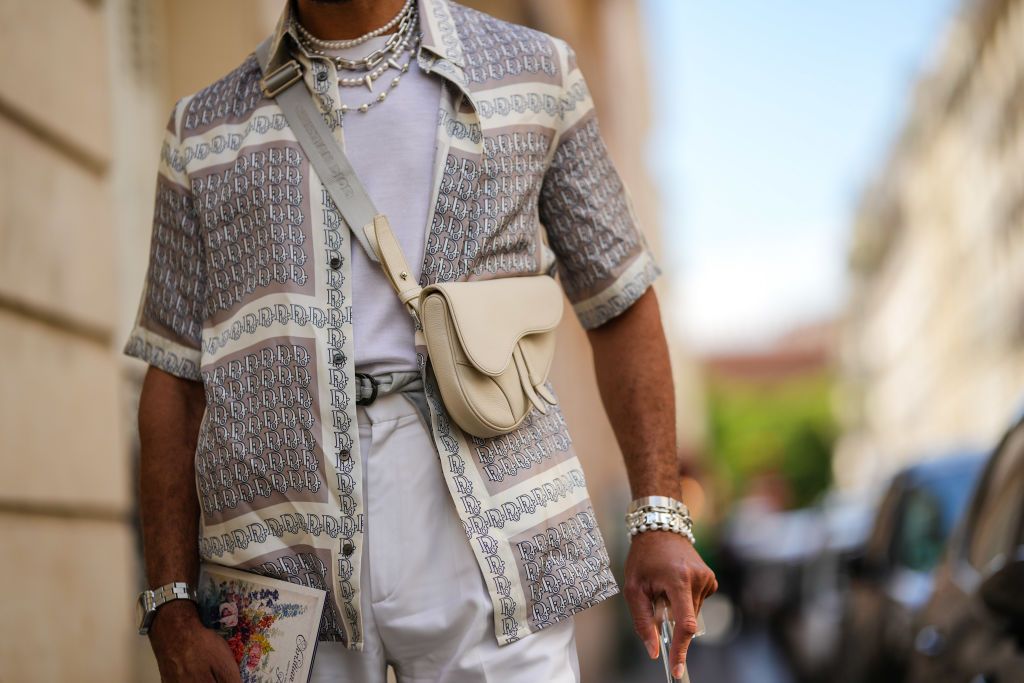 PARIS, FRANCE - JUNE 24: A guest wears a white high neck t-shirt, a gray and white latte print pattern short sleeves / silk shirt from Dior, a beige matte leather crossbody Saddle bag from Dior, a silver chain bracelets, a silver watch, silver rings, white suit pants, a silver crutch, outside the Dior Homme show, during Paris Fashion Week - Menswear Spring/Summer 2023, on June 24, 2022 in Paris, France. (Photo by Edward Berthelot/Getty Images)