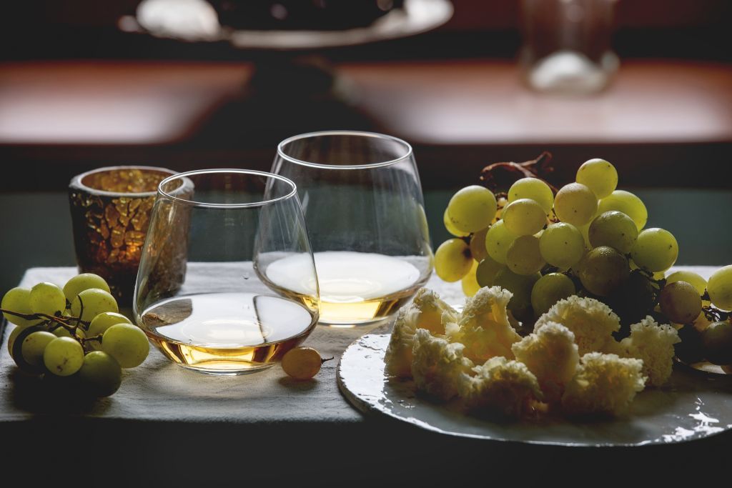Swiss cheese tete de moine on ceramic board, black and green grapes and two glasses of white wine, served with candle on white table cloth with window behind. Natural day light. Romantic dinner.. (Photo by: Natasha Breen/REDA&CO/Universal Images Group via Getty Images)
