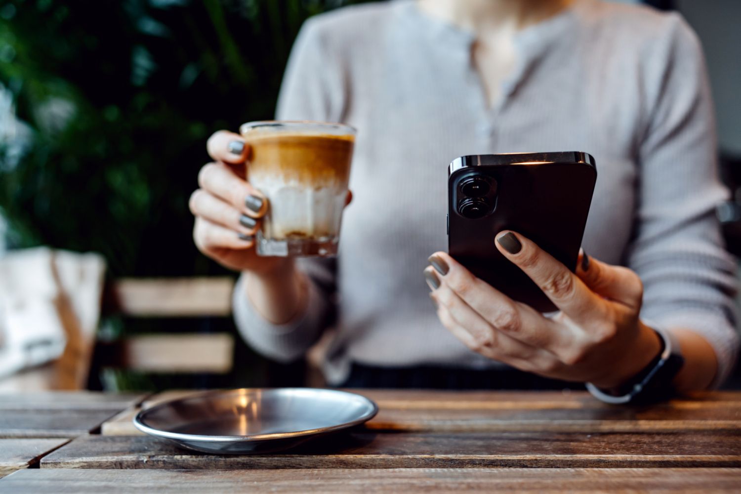 Cropped shot, mid-section of young Asian woman sitting at a table in coffee shop, looking at her smartphone while drinking a cup of coffee. Lifestyle and technology