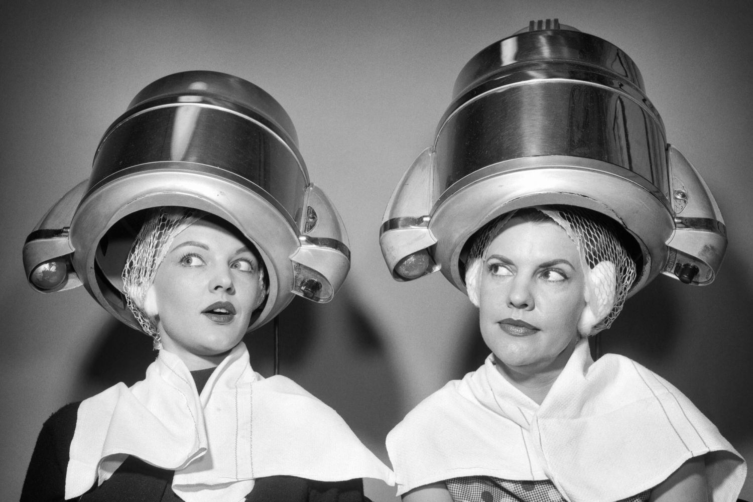 1950s TWO WOMEN SITTING UNDER BEAUTY SALON HAIR DRYERS WEARING HAIRNETS TOWELS TALKING GOSSIP (Photo by Debrocke/ClassicStock/Getty Images)