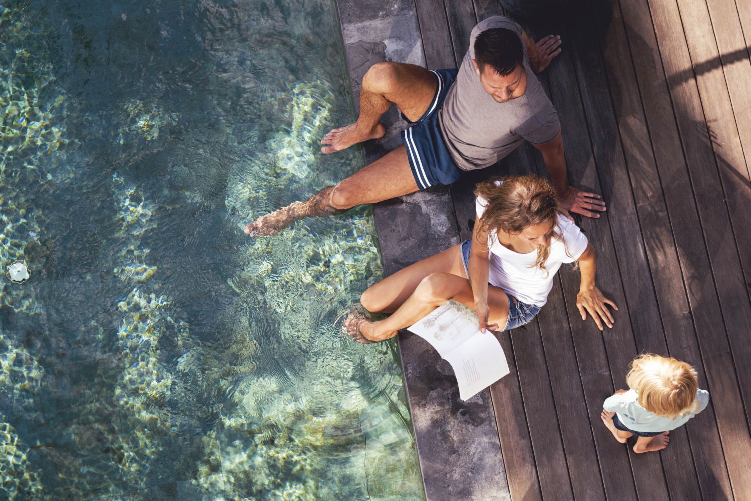 birds view of parents sitting next to pool while mother is holding a book and speaking with her young son