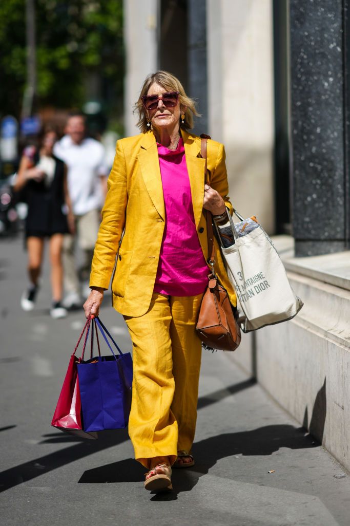 PARIS, FRANCE - JULY 06: A guest wears pink square sunglasses, gold and pear pendant earrings, gold chain necklaces, a neon pink t-shirt, a yellow mustard blazer jacket, matching yellow mustard flared pants, a brown shiny leather handbag, a white cotton large handbag, a navy blue shopping bag, a burgundy shiny Cartier shopping bag, gold shiny leather belted sandals , outside Viktor & Rolf, during Paris Fashion Week - Haute Couture Fall Winter 2022 2023, on July 06, 2022 in Paris, France. (Photo by Edward Be