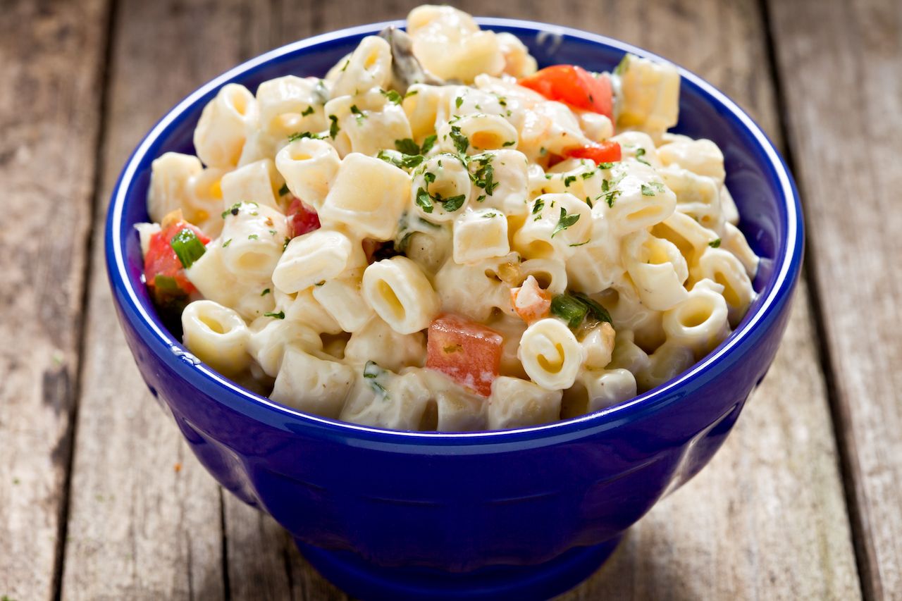 A high angle close up shot of a small blue bowl full of homemade macaroni salad sitting on an old wooden picnic table.