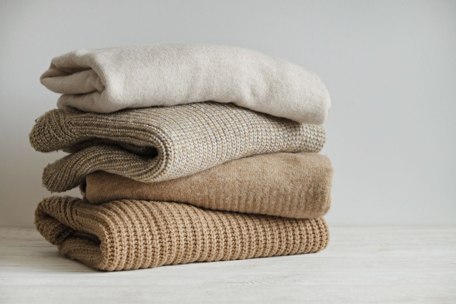 Bunch of knitted warm pastel color sweaters with different knitting patterns stacked in messy pile on white wooden table, white wall background. Fall winter season knitwear. Close up, copy space.