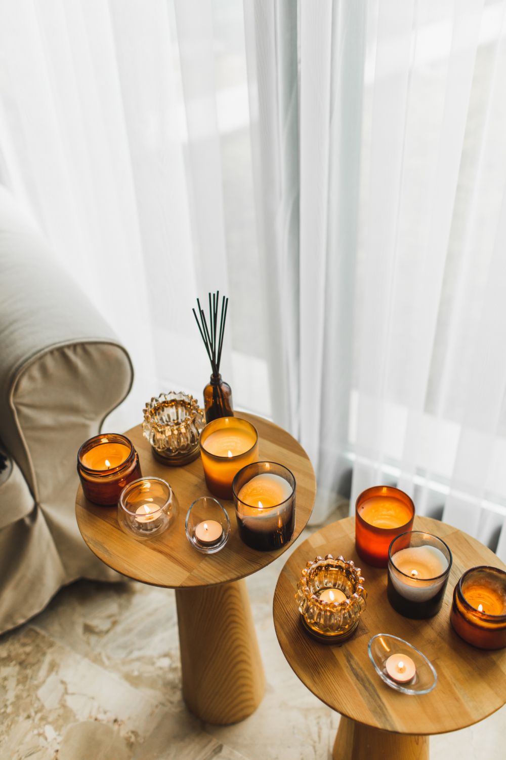 Organic soya wax candles in different glass candlestick in brown bowls on wooden tea-table near comfortable armchair. Cosy, comfortable and soft home background. Hygge and aromatherapy concept.