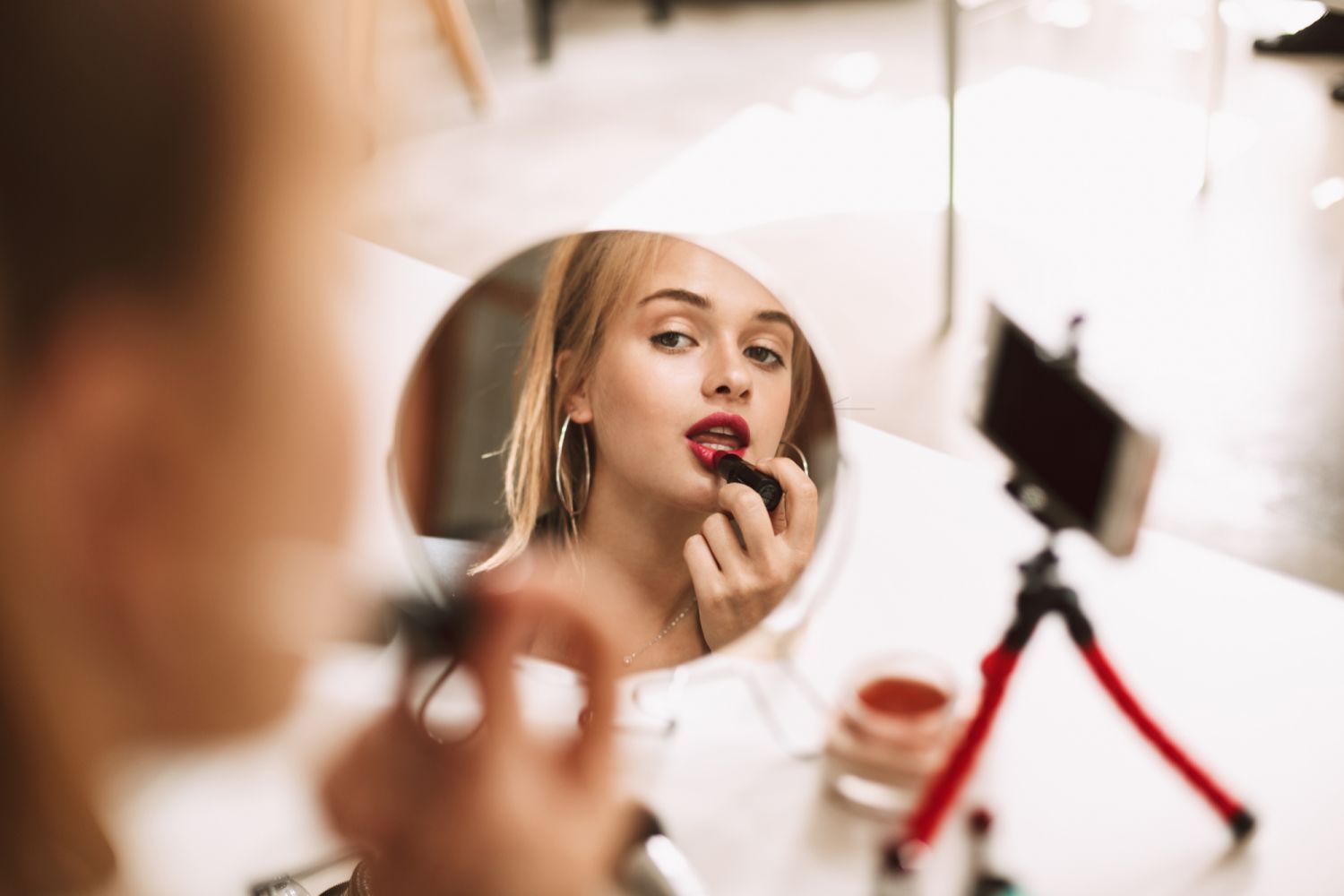 Young pretty girl looking in little mirror on table using red lipstick recording new video for vlog