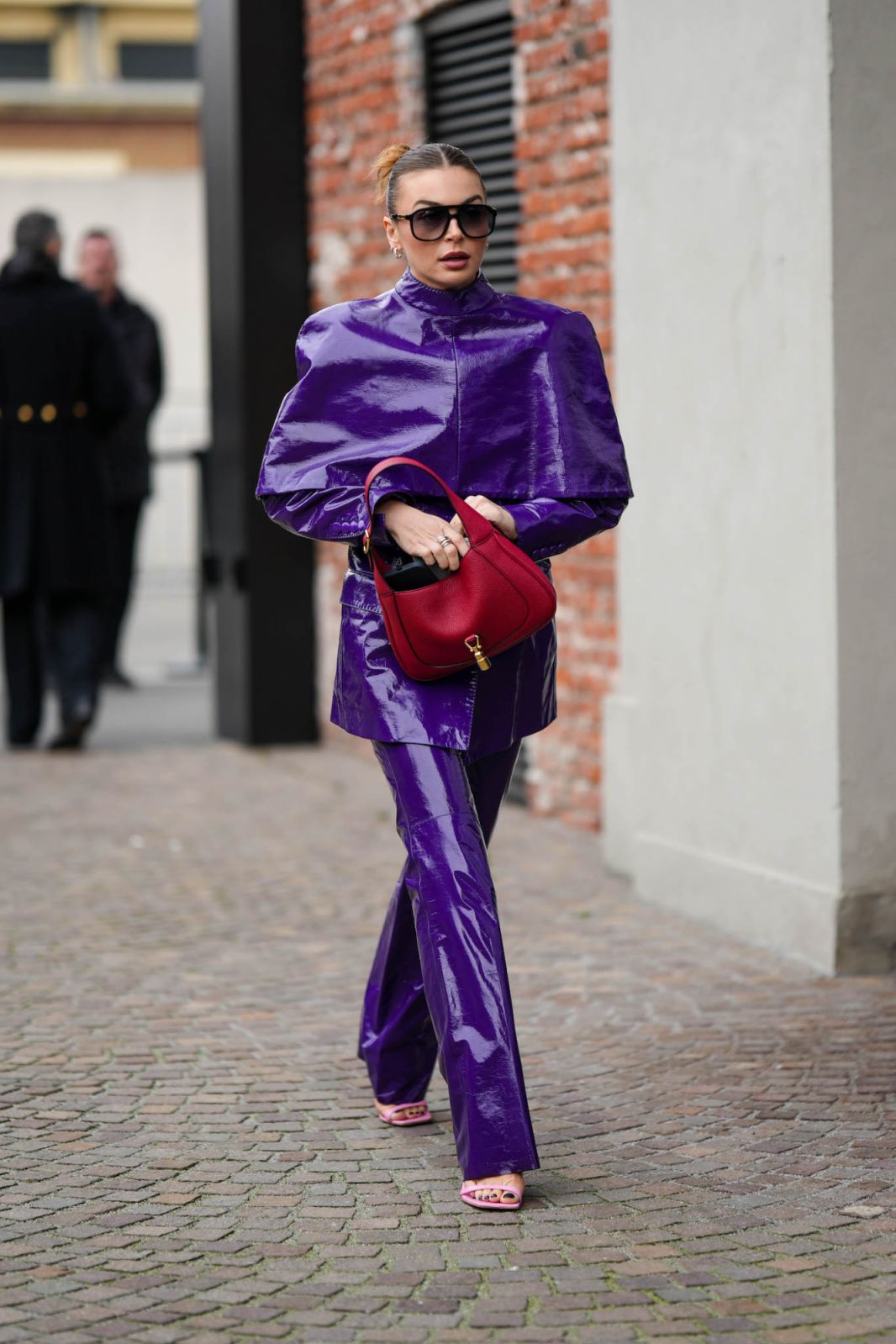 MILAN, ITALY - FEBRUARY 24: A guest wears black sunglasses, a purple shiny varnished leather oversized blazer jacket, neon purple shiny varnished leather large pants, a red shiny leather handbag from Gucci, pink shiny leather open toe-cap heels mules , outside Gucci, during the Milan Fashion Week Womenswear Fall/Winter 2023/2024 on February 24, 2023 in Milan, Italy. (Photo by Edward Berthelot/Getty Images)